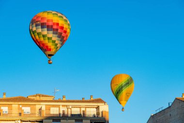 Guadix, Granada - Spain - 01-24-2024: Two hot air balloons soar above houses with red roofs; one is a colorful checked balloon and the other is yellow. clipart