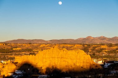 Guadix, Granada - İspanya - 01-24-2024: Guadix 'teki bir kaya oluşumuna geniş bir bakış açısı, batan güneşle aydınlanıyor, mavi gökyüzünde ay var..