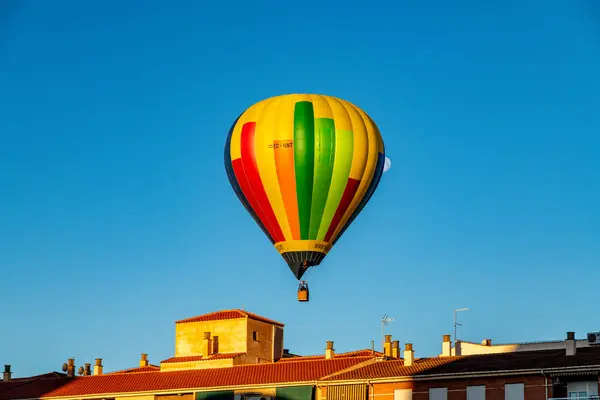 Guadix, Granada - İspanya - 01-24-2024: Sarı, yeşil ve kırmızı renkli canlı bir sıcak hava balonu açık mavi gökyüzüne kırmızı çatıları olan evlerin üzerinde yükseliyor,