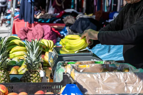 Guadix, Granada - İspanya - 01-24-2024: Ön planda ananas gibi çeşitli meyvelerle muz tartan bir satıcının yakın çekimi.