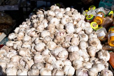 Guadix, Granada - Spain - 01-24-2024: A large pile of fresh garlic bulbs on a market stall, surrounded by jars of pickled products shining in the sunlight. clipart