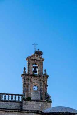 Close-up of a church bell tower with a clock and stork nest, showcasing stone details and a blue sky backdrop.