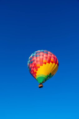 A Balloons hot air balloon soars in the clear blue sky, its vibrant checkered envelope in red, yellow, blue, and green, evoking adventure. clipart