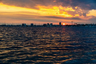 Güneş sakin bir kıyı koyunun üzerinde batıyor. Gökyüzünde canlı turuncu ve mor renkler bırakıyor. Siluetli binalar ve palmiyelerle..