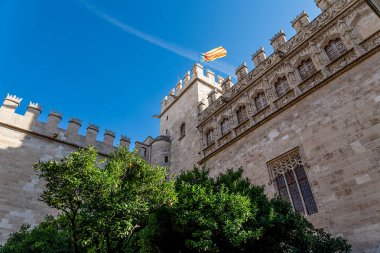 Siperleri ve kulesi olan tarihi bir cephe. Arka planda Gotik detaylar ve süslemelerin yer aldığı İspanyol bayrağı..