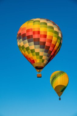 Açık mavi gökyüzüne yükselen iki renkli sıcak hava balonu. Biri canlı bir kareli desen içerirken, diğeri sarı-yeşil çizgili..