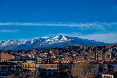 Guadix, Granada - İspanya - 01-24-2024: Guadix şehri, engebeli bir kaya manzarası ve açık mavi gökyüzü altında karla kaplı dağlar tarafından çerçevelenmiş önplandaki kentsel binaları sergiliyor.