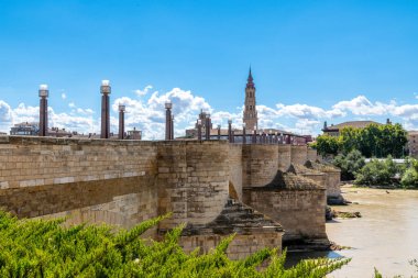 Büyük sütunları olan taş bir köprü, modern ışık direkleri, bir şehir silueti ve parçalı bulutlu bir gökyüzünün altında kendine özgü bir kule..