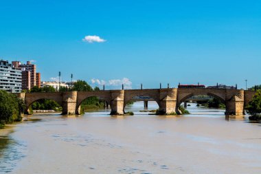 Puente de Piedra Ebro nehri üzerinde kemerler ve sokak lambaları, arka planda modern binalar ve açık mavi gökyüzü..