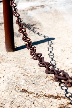 Kalın, paslı bir zincir, metal bir direğe bağlı, kumlu zeminde çaprazlama ilerliyor. Geniş, yıpranmış halkalar gölgeler oluşturarak ağırlığını ve dokusunu arttırır..