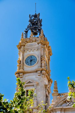 Valencia Belediye Binası 'nın saat kulesi, bir çan ile süslenmiş ve yeşil yapraklarla çerçevelenmiş, parlak mavi bir gökyüzüne karşı duruyor..