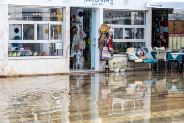 Tavira, Algarve - Portekiz - 09-18-2024: Dükkan camları, renkli seramik figürler, el yapımı çantalar ve bir kafe ile dolu sel basmış bir cadde.