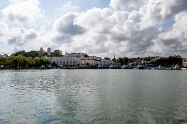 Tavira, Algarve - Portekiz - 09-18-2024: Yüksek su seviyesi, tarihi binalar ve bir köprüye sahip bir nehrin panoramik manzarası. Nehir kıyısında ağaçlar var. Parçalı bulutlu gökyüzü.