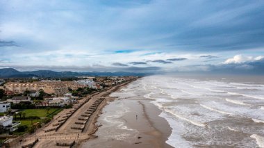 Denia, Alicante - İspanya - 11-13-2024: Yumuşak dalgalar, kum tepeleri ve suda yürüyen bir insanın bulunduğu uzun kıyı şeridi.