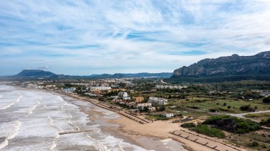 Denia, Alicante - İspanya - 11-13-2024: Kum sahili boyunca dalgaların kırıldığı ve arka planda dağların olduğu kıyı manzarası.