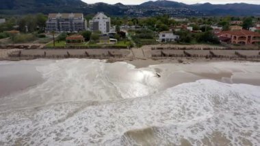 Denia, Alicante - İspanya - 11-13-2024: İnsansız hava aracı sahilde soldan sağa uçuyor, dalgaları, kumulları, evleri, dağları ve arka planda karanlık, bulutlu gökyüzünü gösteriyor.