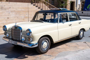 Saragossa, Saragossa - Spain - 09-10-2021: Front view of a cream-colored Mercedes-Benz 200D classic, parked on paved area, with chrome grill, round headlights, and modern buildings. clipart