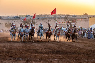 Agadir, Souss-Massa - Fas - 08-03-2024: Geleneksel giysiler içindeki berber sürücüler, günbatımında bayraklar tutarak tüfeklerle süslü atlar üzerinde Fantasia sergilediler.