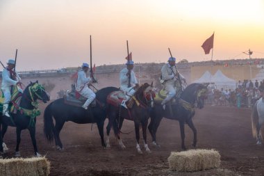 Agadir, Souss-Massa - Fas - 08-03-2024: Süslü atlar üzerindeki üç Berber binicisi tüfekleri tutuyor ve arka planda seyircilerin yer aldığı gün batımında Fantasia 'nın gösterisini bekliyorlar.