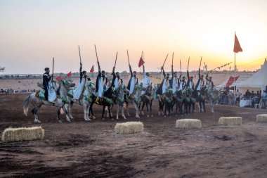 Agadir, Souss-Massa - Fas - 08-03-2024: Berber sürücüler tüfeklerle süslenmiş atların üzerinde dizilmiş vaziyetteler, gün batımında arka planda sallanan bayraklar, neşeli bir atmosfer yaratıyor.