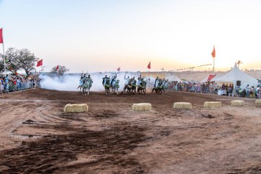Agadir, Souss-Massa - Fas - 08-03-2024: Geleneksel giyinmiş Berberi sürücüler arenada Fantasia sergilediler, gün batımında tüfeklerle duman yarattılar, bayraklar sallandı.