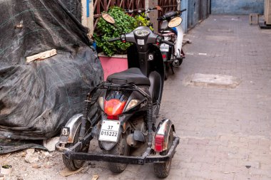Agadir, Souss-Massa - Fas - 08-03-2024: Dar bir sokakta, krom bir bagaj taşıyıcısı ve arka planda yeşil bitkiler bulunan üç tekerlekli siyah bir motosiklet..