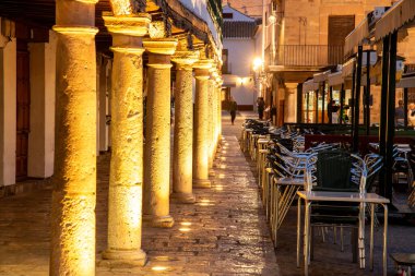 Almagro, Ciudad Real - İspanya - 10-08-2024: Plaza Belediye Başkanı Almagro 'da metal sandalyeler, kaldırımlar ve arka planda akşam atmosferi ile aydınlatılmış sütunların yakın çekimi.