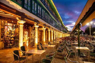 Almagro, Ciudad Real - İspanya - 10-08-2024: Aydınlatılmış atari salonlarıyla Plaza Belediye Başkanı Almagro, tarihi binalar, dükkanlar, açık mobilyalar ve şehir üzerinde gün batımı manzarası.