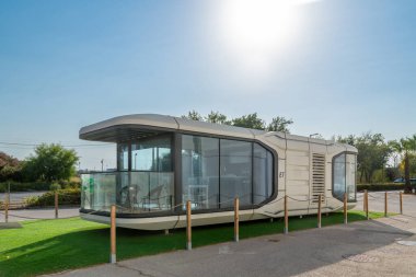 Almancil, Algarve - Portugal - 09-19-2024: Modern modular building with glass front, curved roof, surrounded by wooden posts, path, trees, and sunny sky, creating a futuristic vibe. clipart