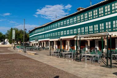 Almagro, Ciudad Real - Spain - 10-09-2024: Traditional building with green wooden windows and white facades at Plaza Mayor, with empty cafe tables under a clear blue sky. clipart