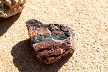 Small angular stone with dark red, orange, and green colors, showing layers and natural unevenness on sandy background. clipart