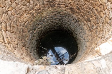 Derin yuvarlak bir taş kuyusu. Mavi gökyüzünü yansıtan su, düzensiz taşın kenarındaki dalları ve bitkileri gösteriyor..