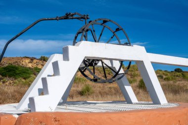 Close-up of a historic water well with a white frame, black wheel, on an orange base, surrounded by dry, grassy land with bushes and hills. clipart