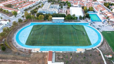 Borba, Alentejo - Portugal - 10-05-2024: Aerial view of a sports complex featuring an artificial turf soccer field, blue track, covered stands, surrounding buildings, and urban area. clipart