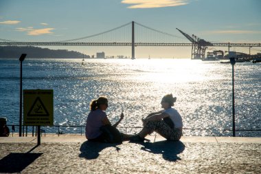 Lissabon, Lisboa - Portekiz - 09-30-2024: İki kadın parlayan Tejo suyu manzaralı bir meydanda oturuyor, Ponte 25 de Abril köprüsü, liman vinci ve bir uyarı işareti.