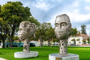 Lissabon, Lisboa - Portekiz - 10-04-2024: Maskeli insan kafası şeklinde iki büyük metal heykel, ağaçlarla çevrili olan Museu de Arte 'de bir Corona anıtı olarak duruyor.