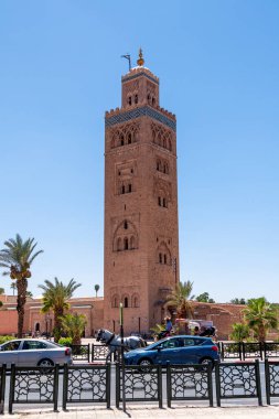 Marrakesch, Marrakesch-Safi - Morocco - 08-14-2024: The minaret of Koutoubia Mosque in Marrakech made of red stone, decorated with ornaments and a golden top, with palms, cars, and a carriage. clipart