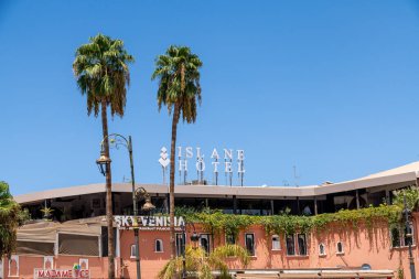 Marrakesch, Marrakesch-Safi - Morocco - 08-14-2024: Islane Hotel in Marrakech with terracotta-colored facade, decorative rooftop sign, surrounded by palms, flowers, and streetlights. clipart