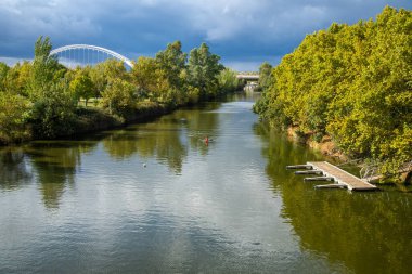 Merida, Badajoz - İspanya - 10-07-2024: Yeşil bankaları olan nehir manzarası, tek bir kanocu ve arka planda çarpıcı beyaz kemerli bir köprü. Bulutlu gökyüzü, sahne aydınlık ve canlı.