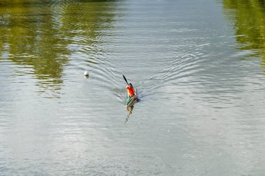 Merida, Badajoz - İspanya - 10-07-2024: Sahil şeridi bitkisinin yeşil yansımalarıyla çevrili kırmızı yelekli tek kanocu. Sahne sakin ve hareketsiz görünüyor..