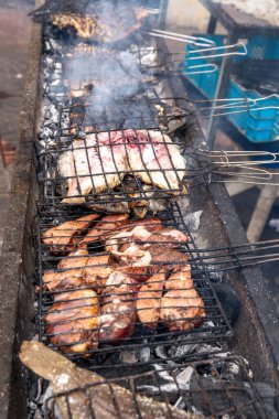 Kömür üzerinde ızgara yapan çeşitli et ve balıklar, pembe balıklar ve tel raflarda altın et parçaları, dumanla çevrili..