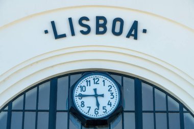 Yuvarlak bir Roma saatine yakın çekim, yukarıda Lisboa metni, modern pencerelerle çevrili ve minimalist detaylarla dolu beyaz bir cephe.