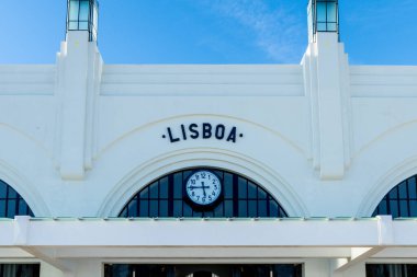 A white building front with large vertical structures, LISBOA in black letters above a clock face, with symmetrical arches and blue sky. clipart