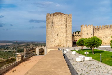 Taş duvarları, kulesi ve tarihi kapısı olan Elvas Kalesi manzarası. Ön planda yeşil alanları ve bankları olan bir kaldırım taşı yolu var..