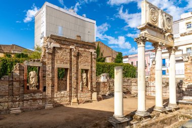 Görüntü, Roma Tiyatrosu 'nun Korint sütunları kısmen restore edilmiş bir bölümünü gösteriyor. Modern binalar ve yeşillik bir karşıtlık yaratır.