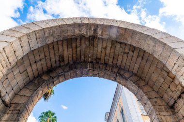 Merida 'daki tarihi bir taş kemerinin yakın plan fotoğrafı. Arka planda palmiyeler ve binalar tarafından çerçevelenmiş devasa taşlar ve gökyüzü manzaralı..