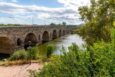 Merida 'daki tarihi Puente Romano, yeşil ağaçlar ve bulutlarla çevrili nehir boyunca uzanan kemerleri olan etkileyici bir taş köprü..