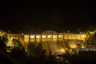 Puerto Pena, Badajoz - Spain - 10-08-2024: A night shot of an imposing dam illuminated with yellow lights, showcasing concrete walls and silhouetted trees below. clipart