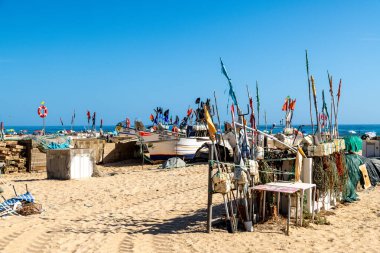 Monte Gordo, Faro - Portugal - 09-14-2024: Fishing boats parked on the beach with flags, nets, and buoys, surrounded by ropes and structures, under a blue sky with tourists. clipart