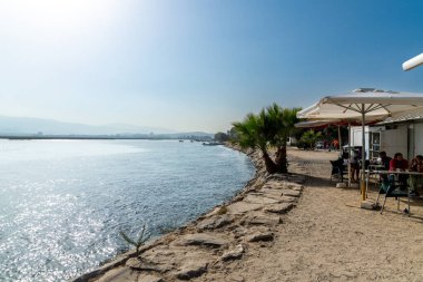 Palmones, Cadiz - İspanya - 08-25-2024: Sakin bir nehir kıyısında, bir restoran ve palmiyelerle nehir kenarı manzarası. İnsanlar şemsiyelerin altında oturur, arkasında dağlar ve gökyüzü vardır..